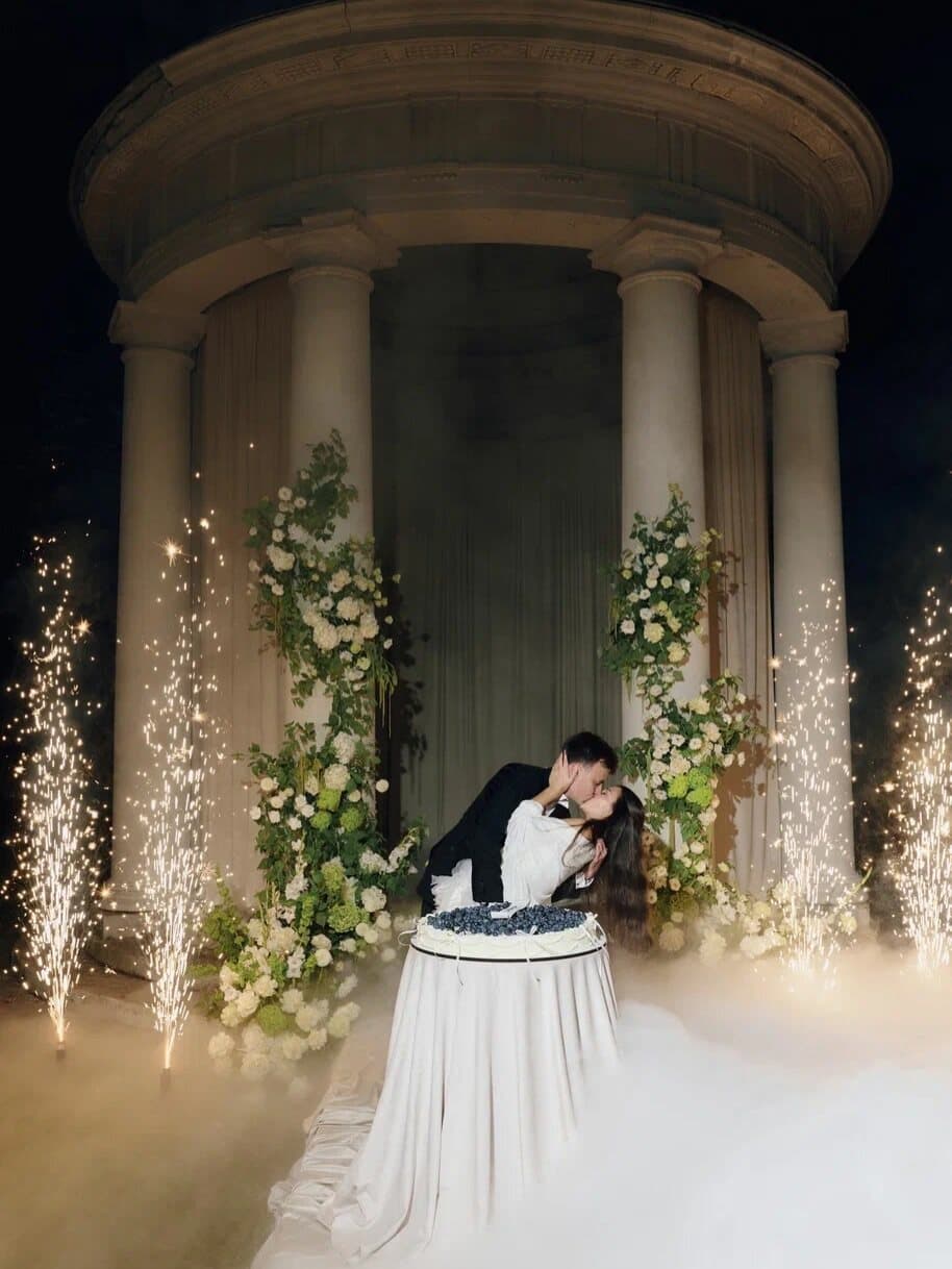 Couple kissing over their cake in front of a columned rotunda, with four cold spark fountains and low fog around them