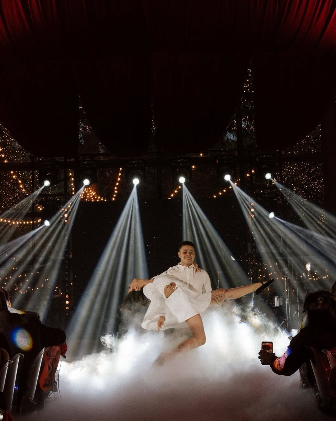 Couple mid-dance in low fog under a row of white moving-head beams while guests film on their phones