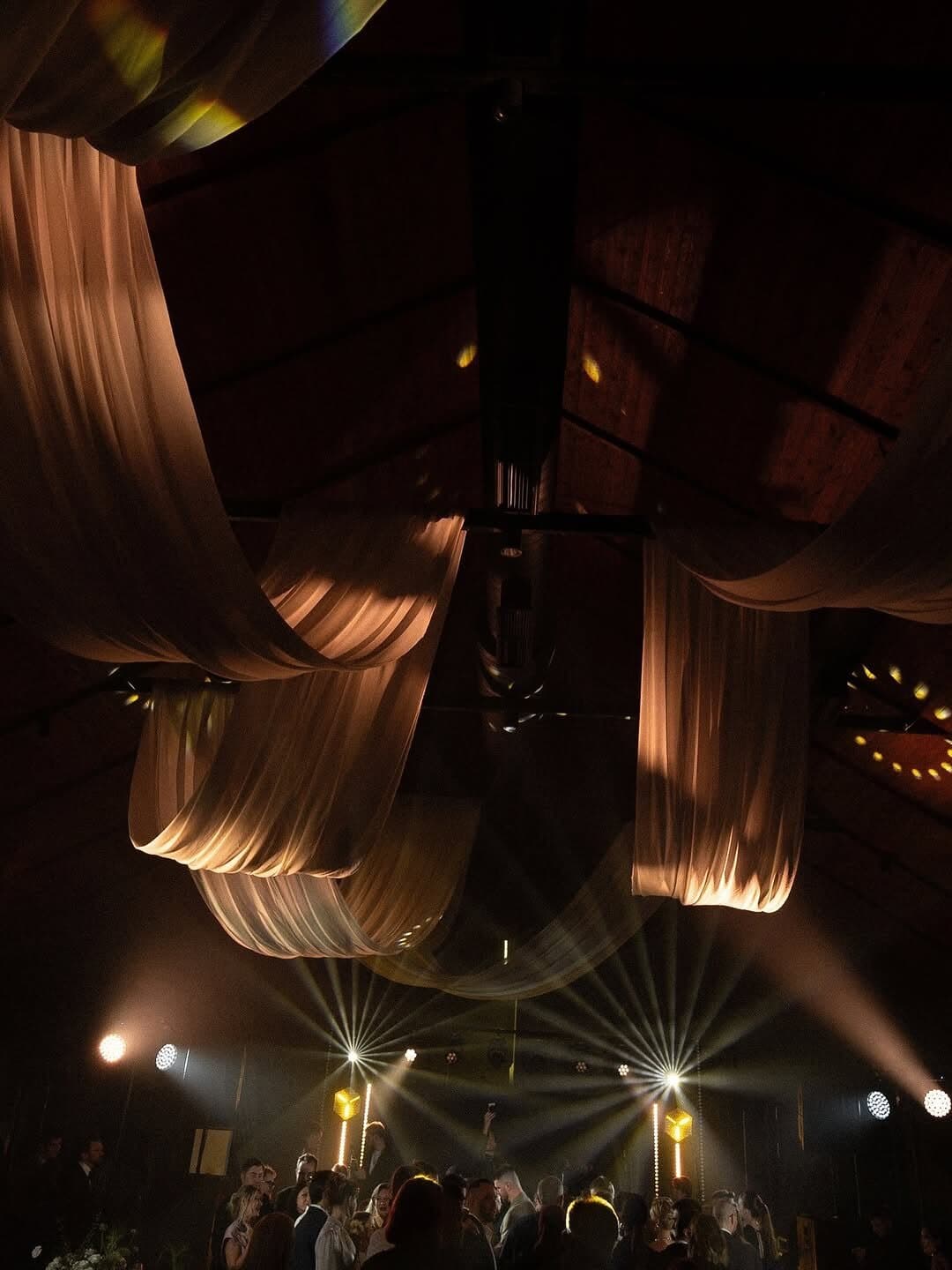 Fabric draped across a reception ceiling glowing warm from uplights, with beams over the dance floor below