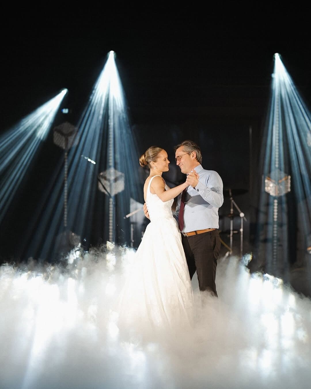 Bride dancing with an older man on low fog under three white light beams