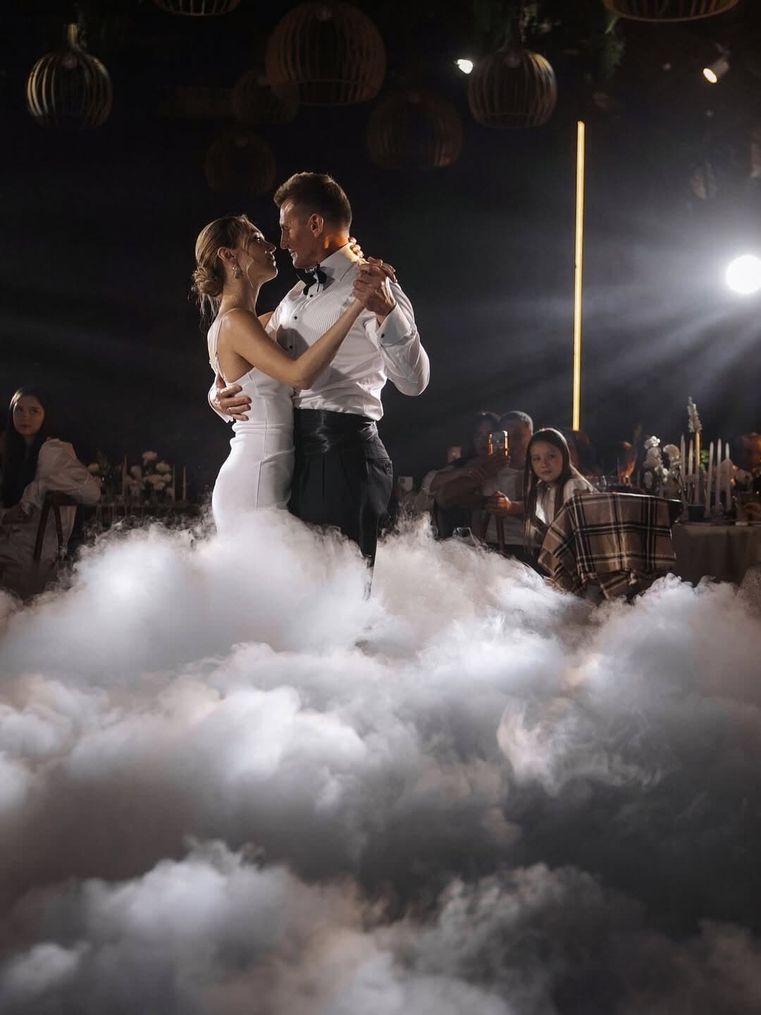 Bride and groom dancing waist-deep in white low fog while guests watch from their tables