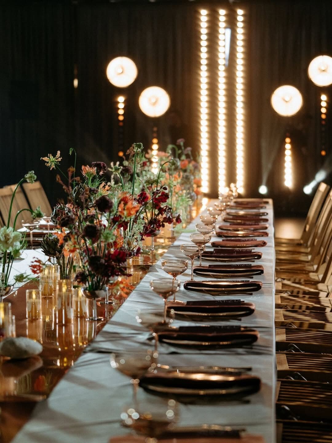 Long candlelit reception table with flowers, framed by warm round lamps and vertical bars of warm bulbs