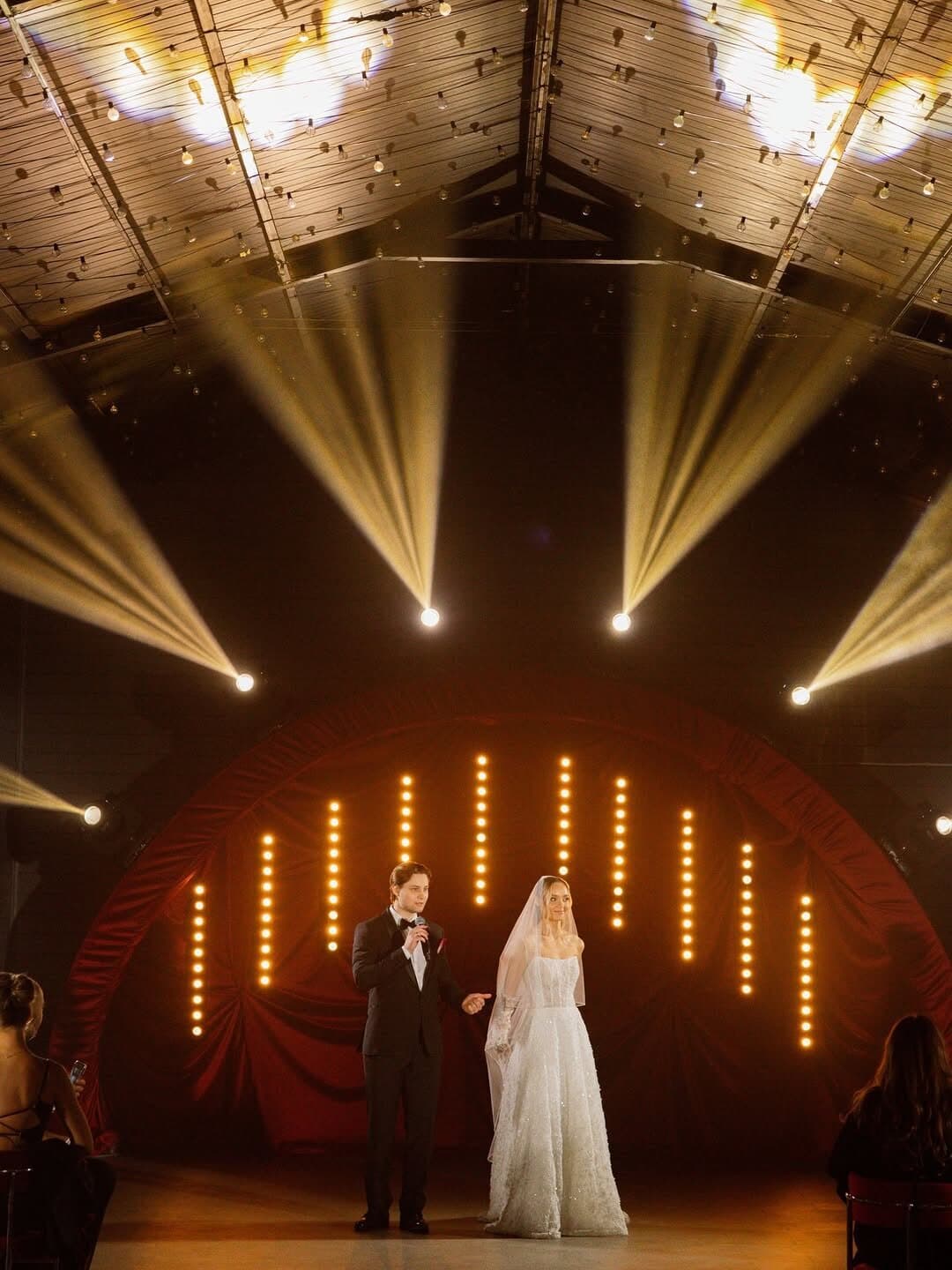 Couple on a small stage giving a toast, with warm beams fanning from the ceiling and vertical warm light bars behind them